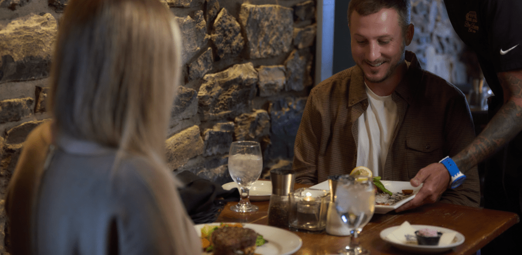 A couple dining at The Tavern as a server delivers their meal, surrounded by the restaurant’s historic stone walls.